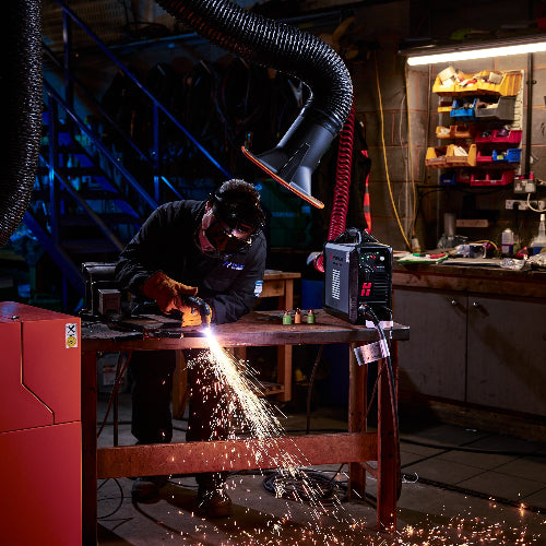 A welder in a dark workshop wearing a welding helmet and gloves, generating bright sparks while plasma cutting metal on a workbench. 